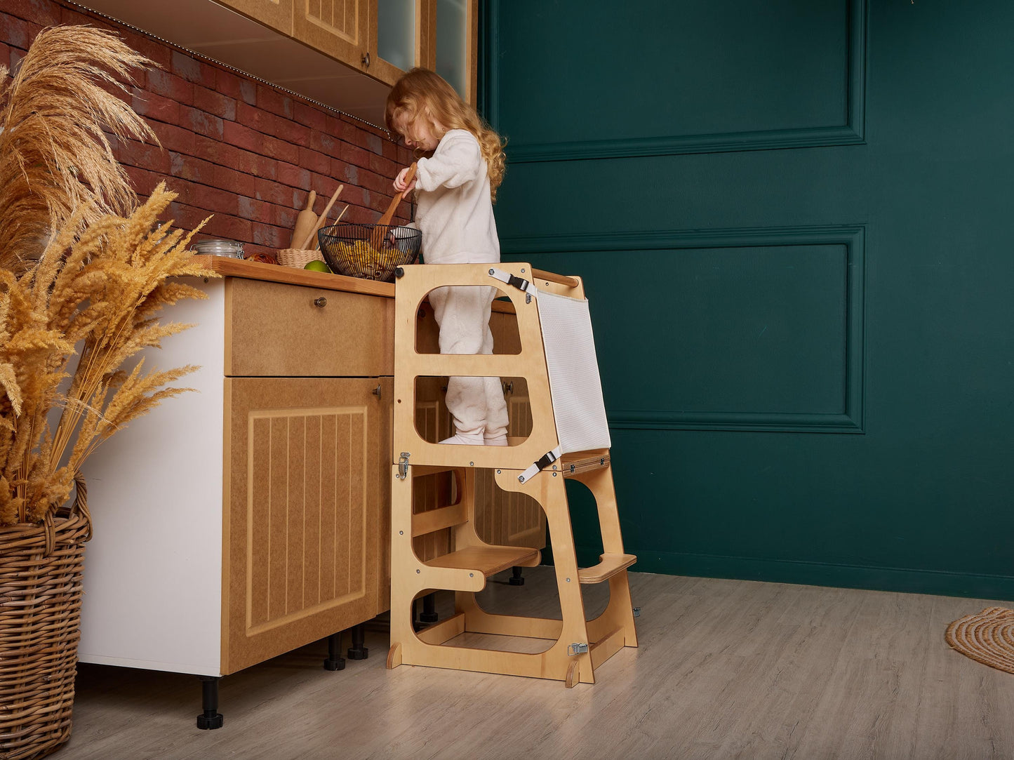 May include: A light wood learning tower with a white canvas panel and a child standing on the top platform. The tower has three steps and a safety rail. The tower is in a kitchen setting with a white cabinet and a brick wall in the background.